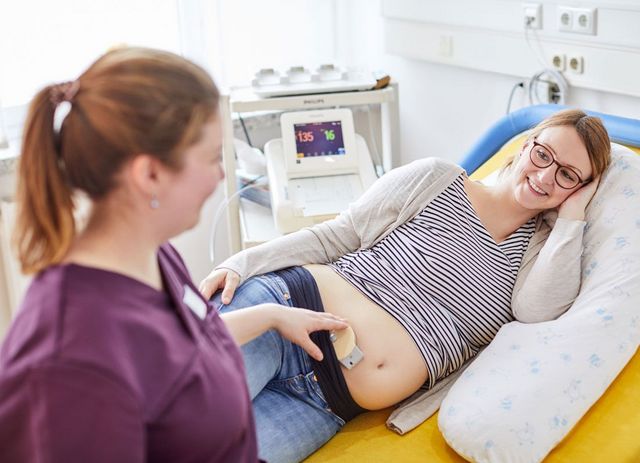 Eine Schwangere liegt seitwärts auf einem Patientenbett. Eine Hebamme untersucht währenddessen den Herzschlag des Babys im Bauch.